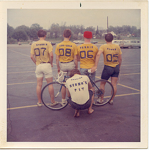 Four guys standing behind a bicycle in a parking lot and one guy kneeling in front. All have their backs to the camera to show the rider numbers and nicknames that are on their shirts.