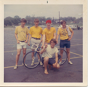 Four high school guys standing behind a bicycle in a parking lot, one guy kneeling in front holding up four fingers. All are geeks and/or minor-level jocks.
